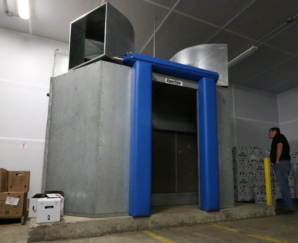 Commercial forced-air cooler with a blue opening beside stacked produce boxes.